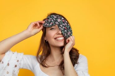 vrouw met oogkussen lachend tegen gele achtergrond.